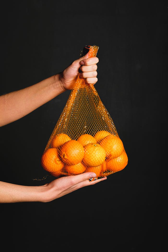 A hand holding a net bag full of fresh oranges against a dark background.